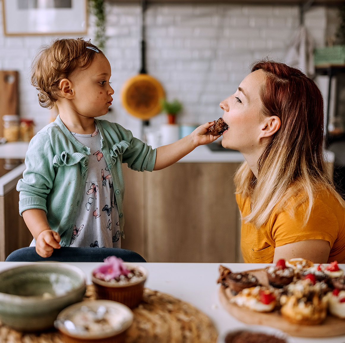 Child feeds woman chocolate treat, both smiling warmly.