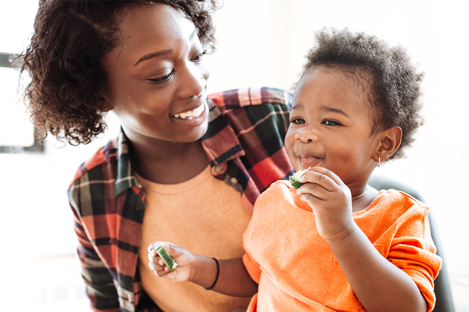 Smiling woman watches toddler eat cucumber slice.
