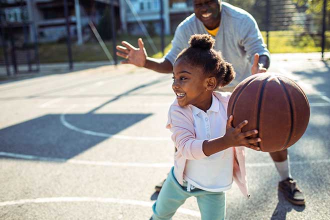 Child playing basketball outdoors, smiling with adult nearby.